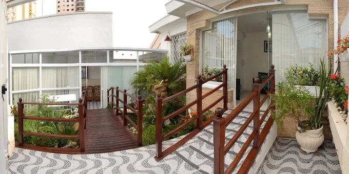 Fachada com rampa de acessibilidade em madeira e corrimão duplo. Entrada de vidro temperado, piso decorado em ondas, pequeno jardim ornamental com plantas e prédios ao fundo no Parque da Mooca.