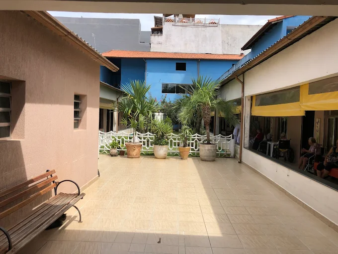 Área externa pavimentada da Casa de Repouso Doce Lar, mostrando um banco de madeira, vasos de plantas ao fundo e janelas onde residentes conversam e socializam.