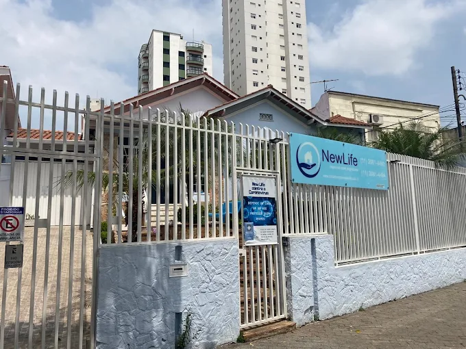 Fachada da Casa de Repouso New Life com muro de pedras azuis, portão e grades brancas. Placa azul com logotipo identifica a unidade e exibe telefones de contato sobre calçada na Mooca.