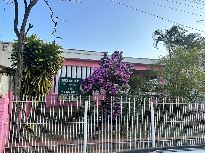 Fachada de cor rosa da Casa de Repouso Protegendo a Vida na Vila Talarico, com grade branca protetora, árvores, plantas e uma grande árvore florida com folhas roxas. Fachada de cor rosa da Casa de Repouso Protegendo a Vida na Vila Talarico, com grade branca protetora, árvores, plantas e uma grande árvore florida com folhas roxas.