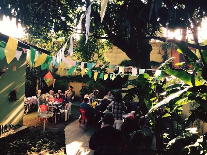 Confraternização de idosos e familiares no jardim arborizado da casa de repouso, decorado com bandeirinhas de Festa Junina. Residentes sentados em mesas sob a sombra de uma grande árvore. Confraternização de idosos e familiares no jardim arborizado da casa de repouso, decorado com bandeirinhas de Festa Junina. Residentes sentados em mesas sob a sombra de uma grande árvore.