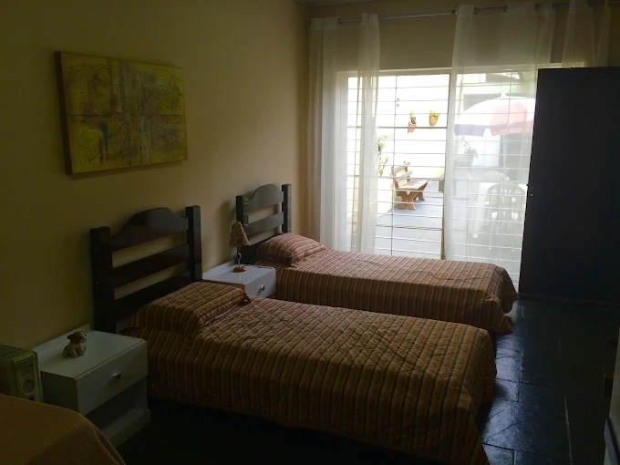 Quarto duplo na clínica geriátrica com duas camas de solteiro organizadas, colchas listradas em tons quentes, criados-mudos brancos e grande janela de vidro com vista para a área externa. Quarto duplo na clínica geriátrica com duas camas de solteiro organizadas, colchas listradas em tons quentes, criados-mudos brancos e grande janela de vidro com vista para a área externa.