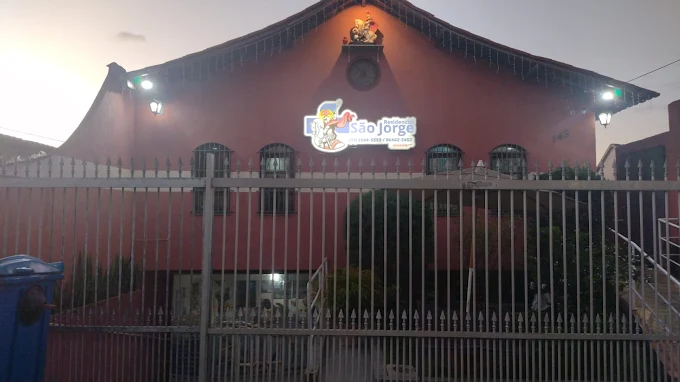Fachada externa da Casa de Repouso São Jorge ao anoitecer. Prédio de tom rosa com portão de ferro alto, janelas gradeadas e o letreiro luminoso da instituição com o telefone visível.