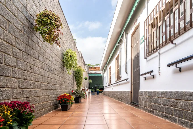 Corredor externo e plano na Shekinah com piso cerâmico, corrimãos duplos de acessibilidade nas paredes, vasos de flores coloridas e plantas suspensas sob céu aberto.