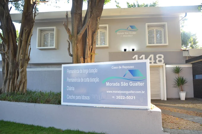 Fachada frontal da Morada São Gualter em Pinheiros. Prédio cinza com grandes árvores e uma placa informativa destacando serviços como longa permanência, diária e creche para idosos.