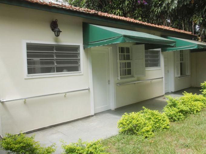Fachada interna térrea do Residencial New Care com barras de apoio metálicas instaladas na parede branca, portas e janelas com toldos verdes e pequeno jardim gramado na frente.