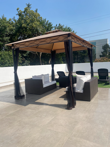 Área externa e aberta da Nostra Casa de Repouso e Spa com piso de porcelanato cinza, gazebo bege com cortinas escuras fluidas, sofás de fibra sintética marrom com estofado branco e espreguiçadeiras ao fundo sob céu azul. Área externa e aberta da Nostra Casa de Repouso e Spa com piso de porcelanato cinza, gazebo bege com cortinas escuras fluidas, sofás de fibra sintética marrom com estofado branco e espreguiçadeiras ao fundo sob céu azul.