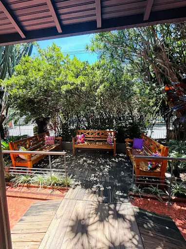 átio externo sombreado por árvores no Residencial Mais Vida na Vila Mariana. Três bancos de madeira com almofadas coloridas circundam o chão de blocos salpicado por pequenas flores caídas. átio externo sombreado por árvores no Residencial Mais Vida na Vila Mariana. Três bancos de madeira com almofadas coloridas circundam o chão de blocos salpicado por pequenas flores caídas.