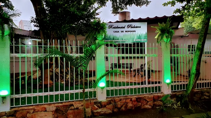Fachada do Residencial Pinheiros à noite, destacando grade de proteção branca iluminada por luminárias verdes. Há palmeiras na entrada e uma placa com o logotipo da casa de repouso e o telefone (11) 3021-5077 bem visível.