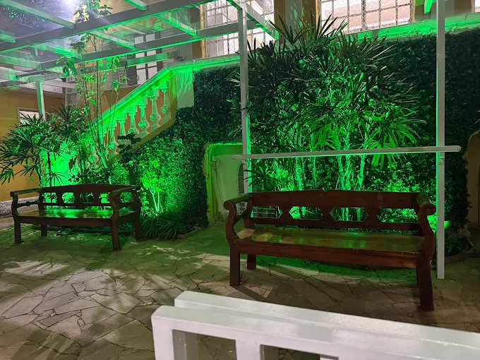 Pátio externo com piso de pedra da VIP SENIOR RESIDENCIAL à noite. Dois bancos de madeira estão posicionados sob uma estrutura de vidro com plantas e folhagens iluminadas por luzes verdes. Pátio externo com piso de pedra da VIP SENIOR RESIDENCIAL à noite. Dois bancos de madeira estão posicionados sob uma estrutura de vidro com plantas e folhagens iluminadas por luzes verdes.